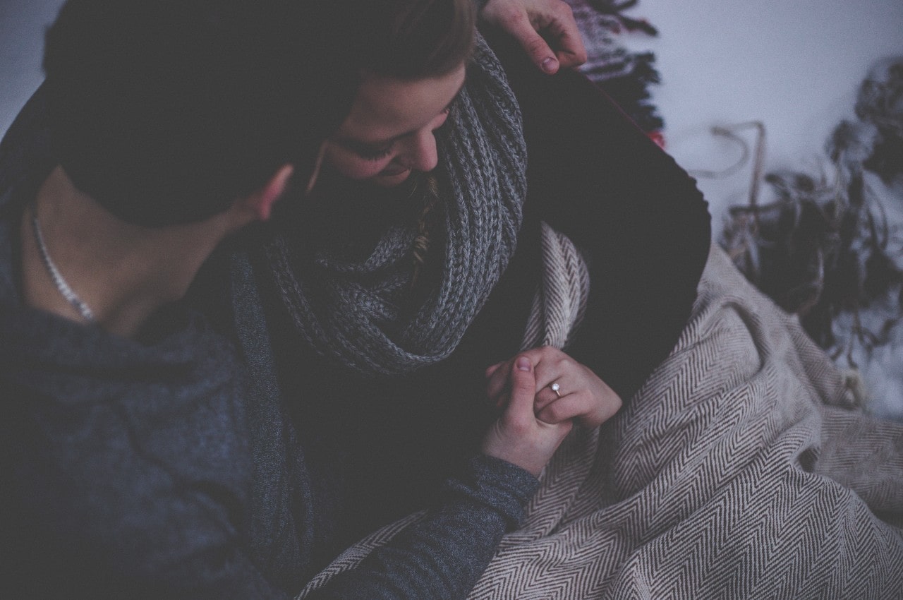 A couple huddled together under a blanket, the woman wearing a diamond engagement ring