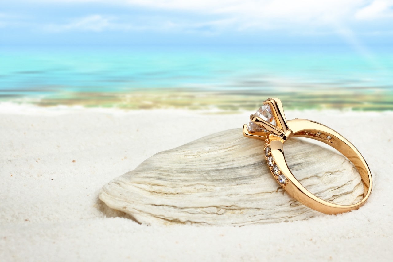 A gold sidestone engagement ring leaning on a seashell by the seashore