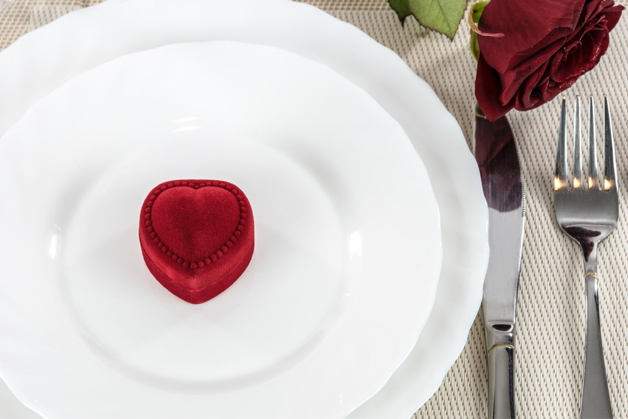 A small, heart shaped box on a white dinner plate at a table setting with a fork and knife next to the plate along with a red rose