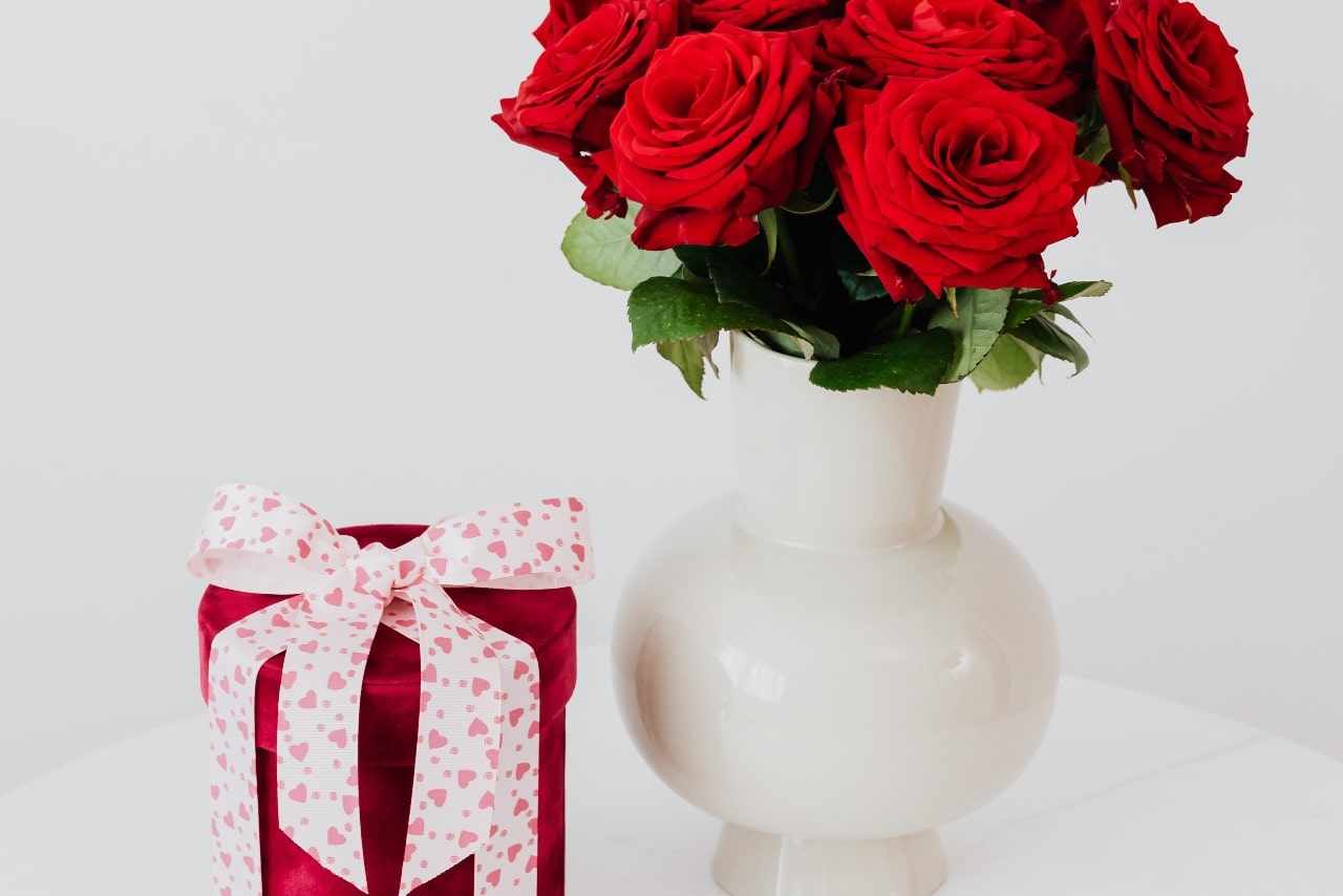 A bouquet of red roses in a white vase next to a cylinder velvet box with a white and pink heart bow on top