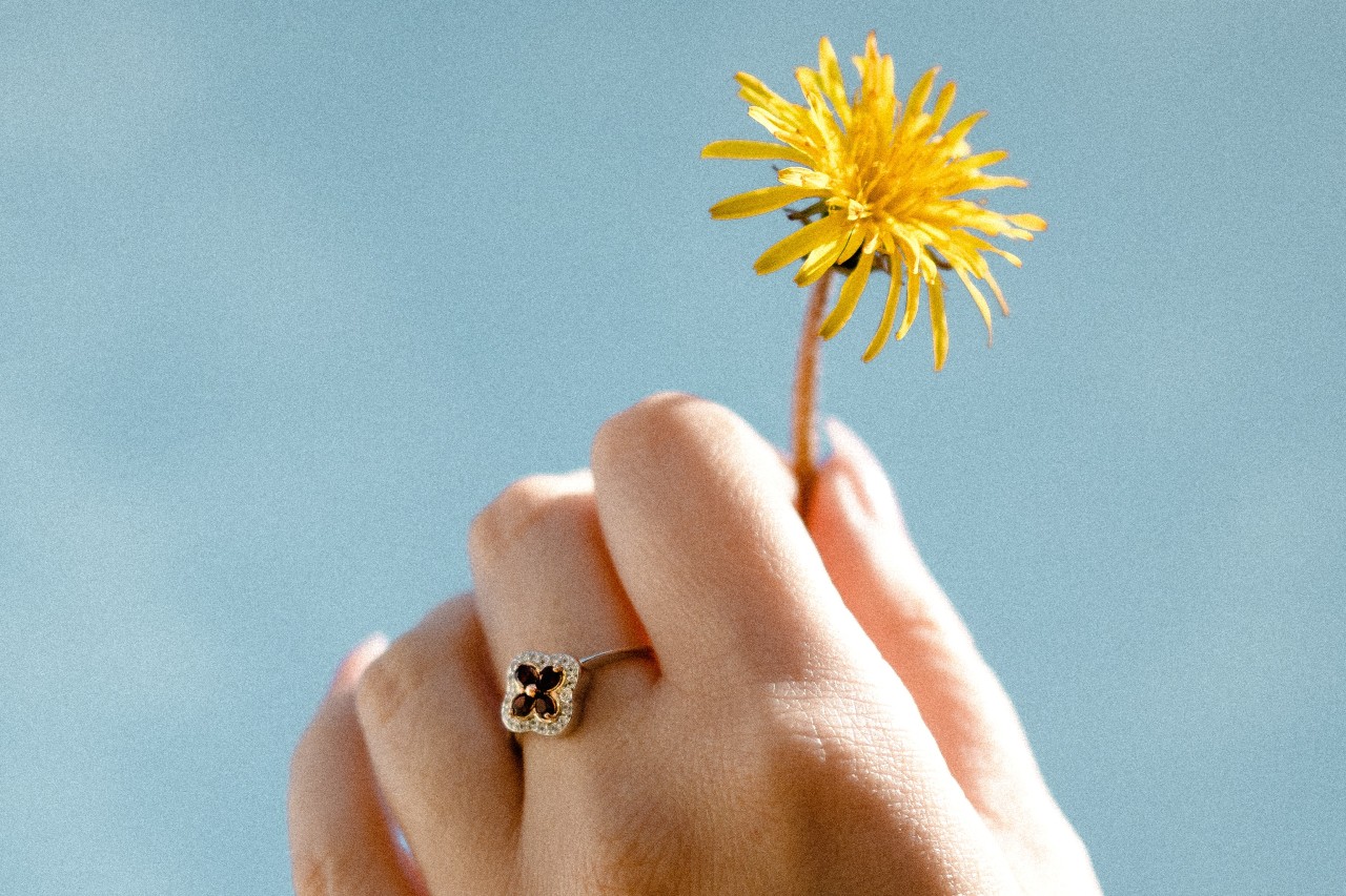 A hand holding a dandelion and wearing a stylized floral ring with red gemstones