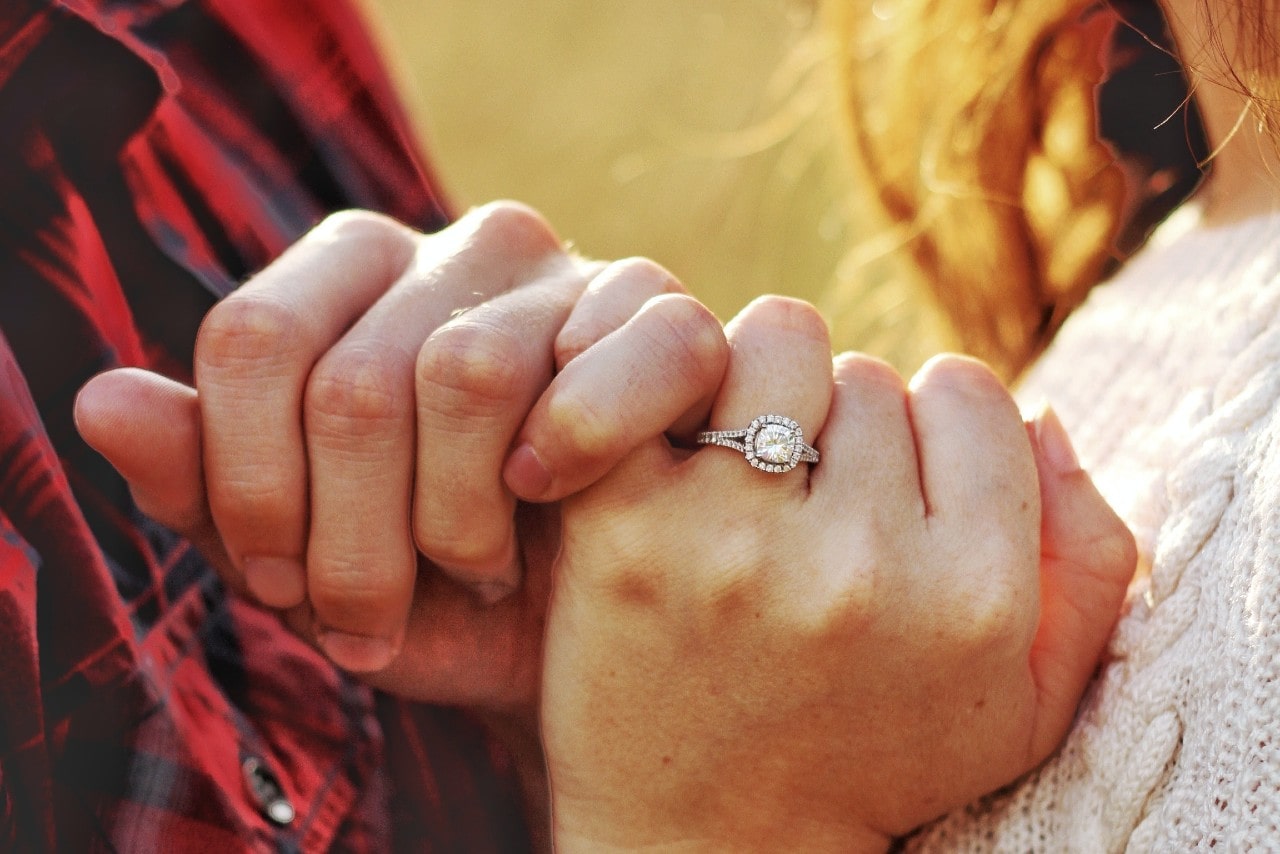 A couple intertwining pinkies, the woman wearing a silver halo engagement ring