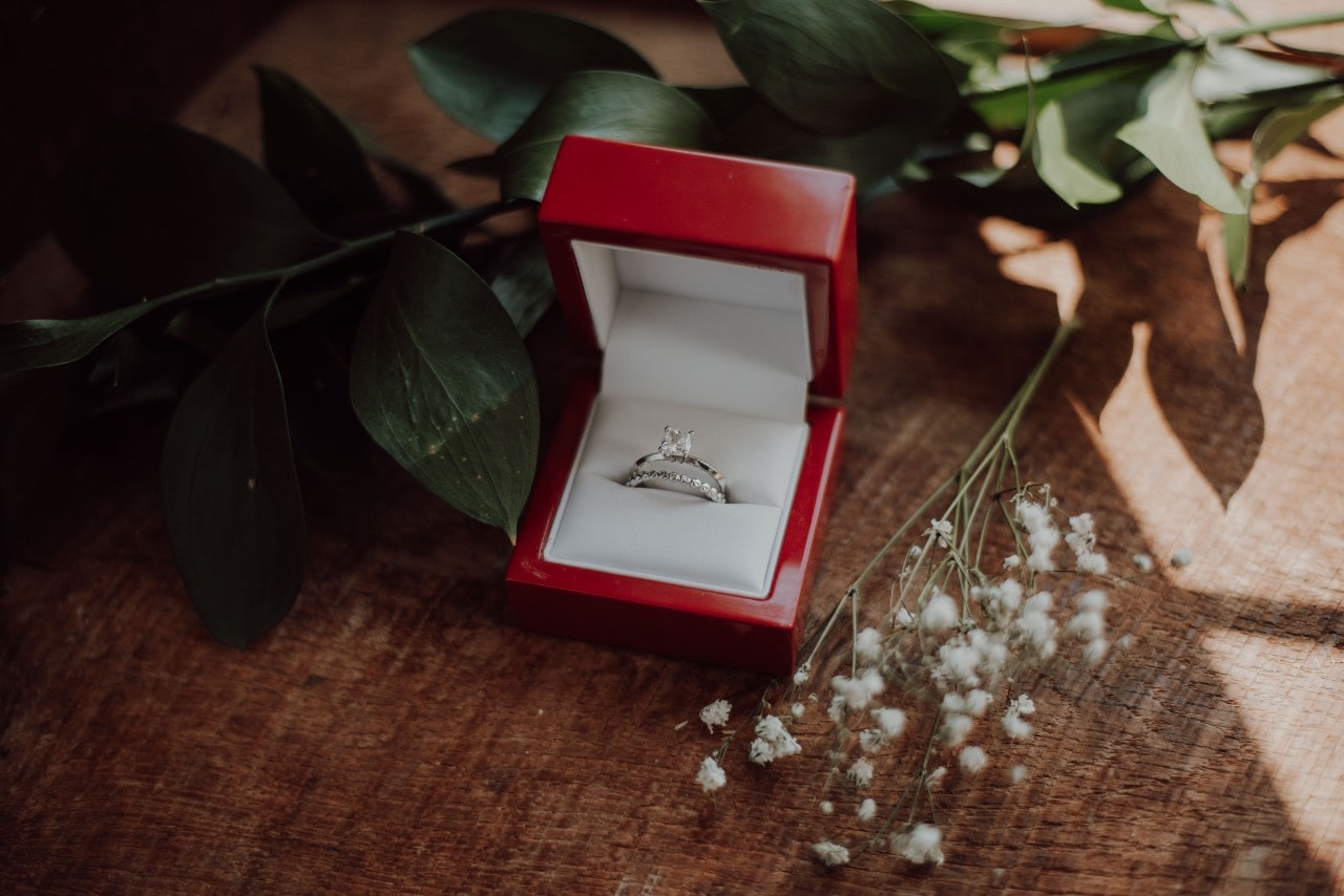 An engagement ring and wedding band sit in a red ring box with flowers