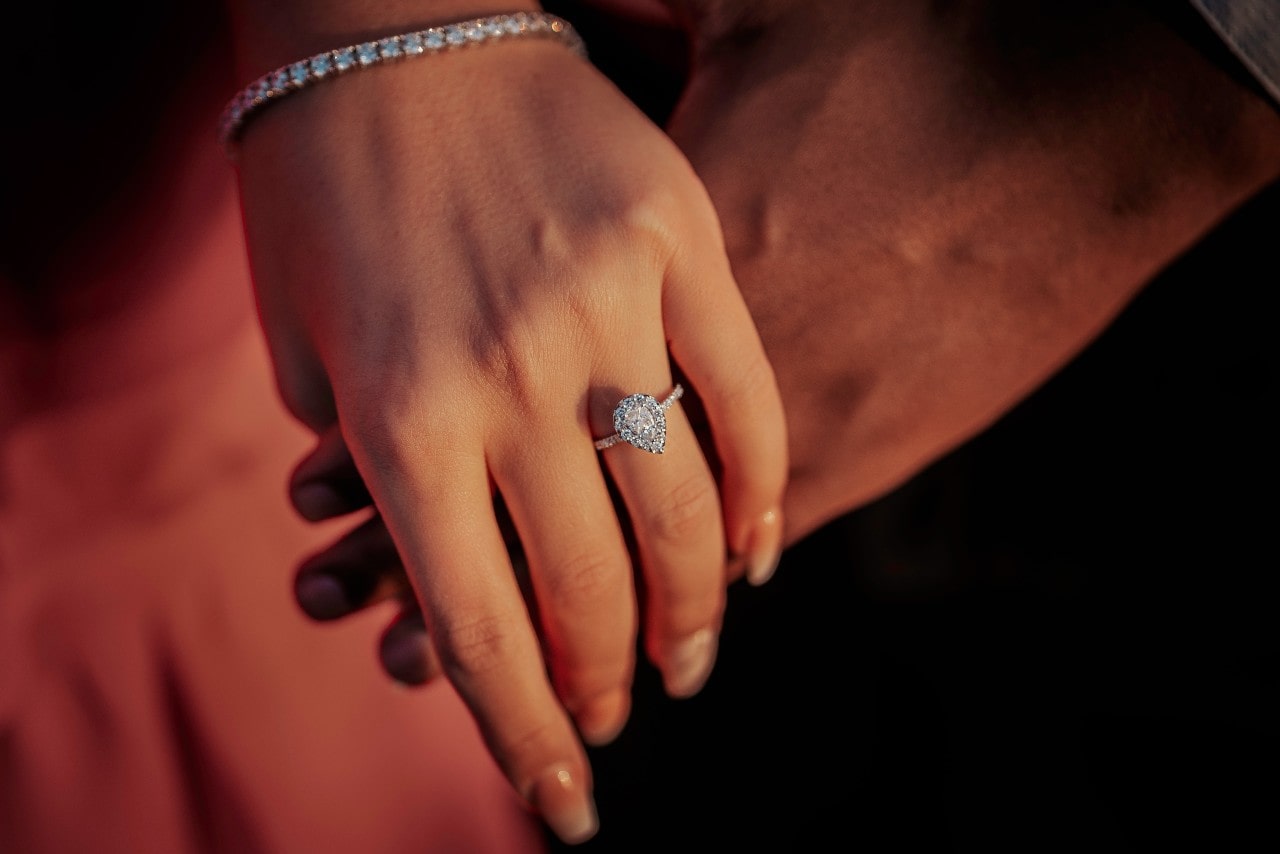 Close up image of a woman’s hand resting on top of her love’s hand showing her pear shape halo engagement ring with diamond bracelet
