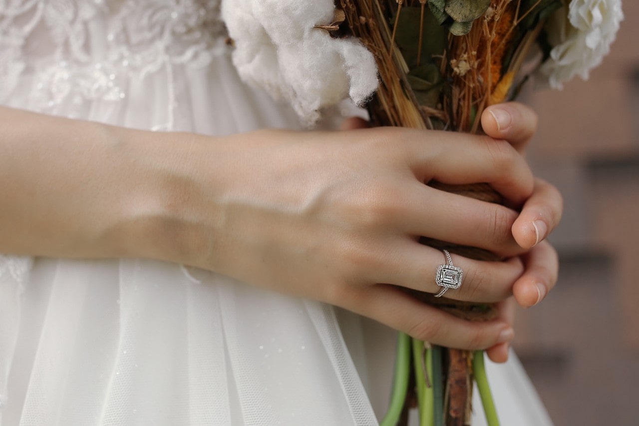 A bride holding a bouquet of flowers wearing an emerald cut diamond engagement ring with a halo and diamonds along the band