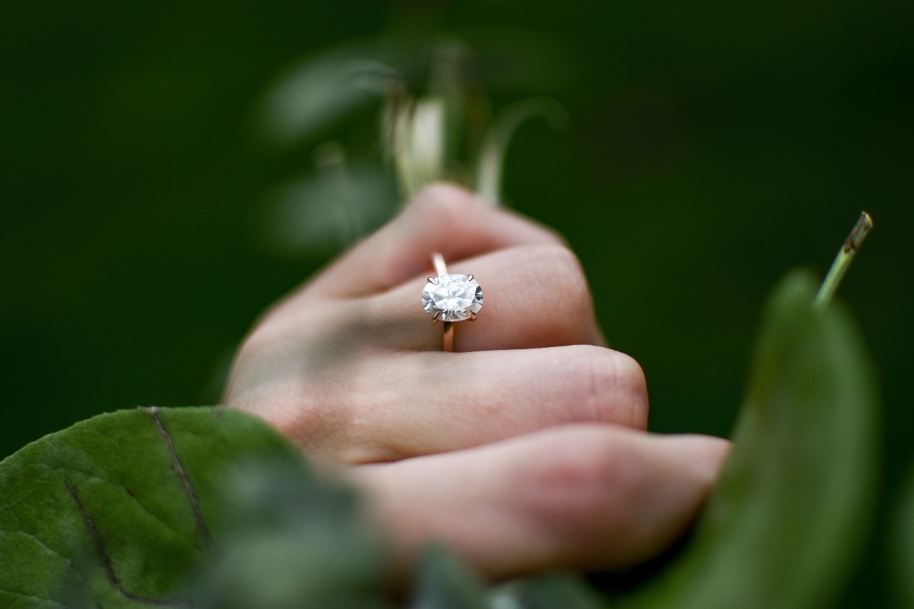 A person’s hand wearing a solitaire, oval cut engagement ring surrounded by greenery