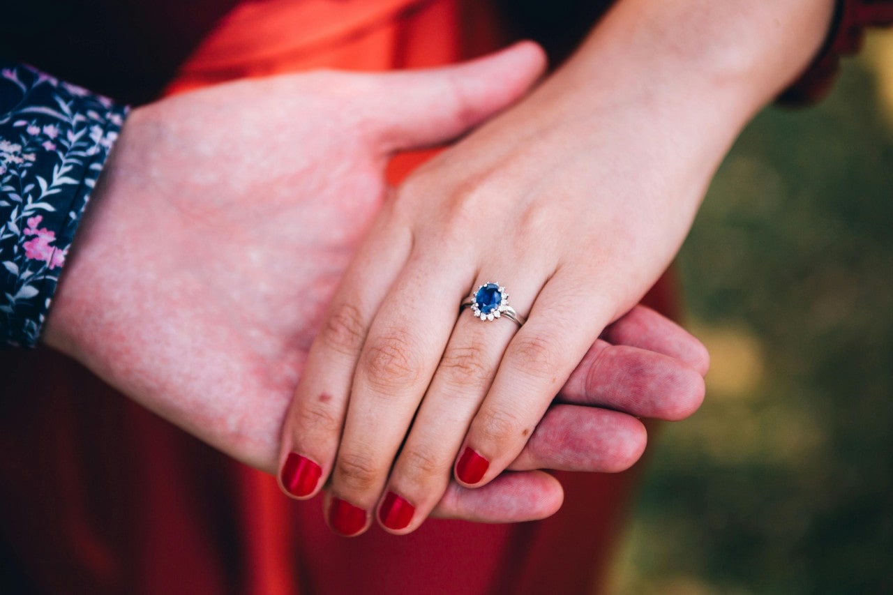 A close-up of a man’s hand holding a woman’s hand that is adorned with a sapphire halo engagement ring.