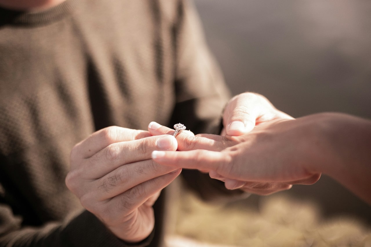 A man sliding an engagement ring onto a woman’s ring finger.