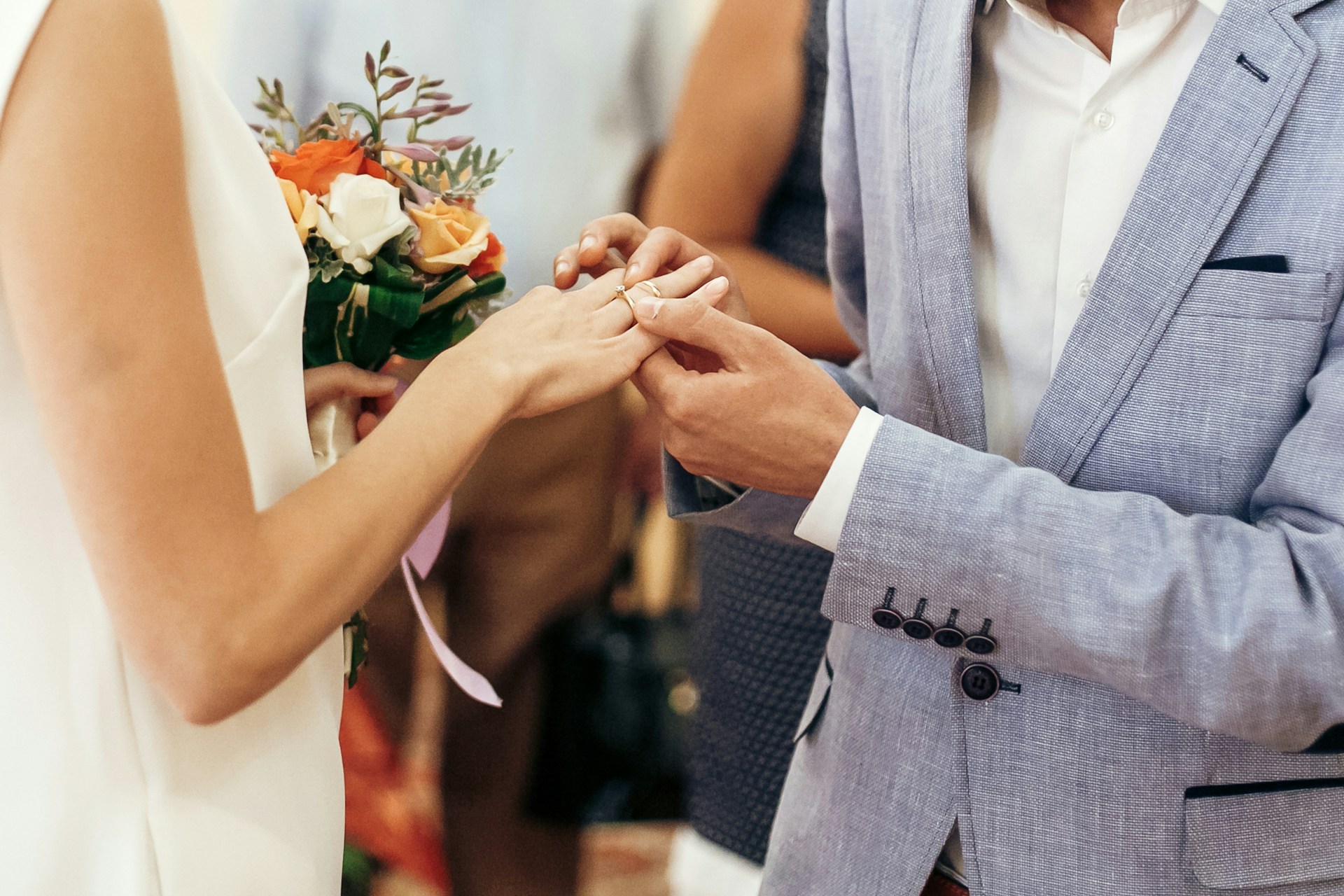 A close-up of a groom slipping a wedding band on his bride’s finger as they stand before a crowd at the altar.