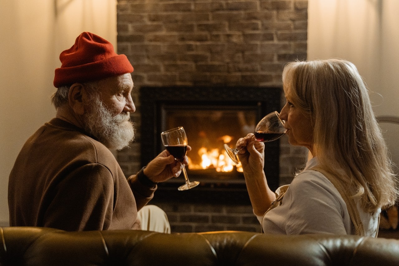 An older married couple sits by the fireplace and sips red wine.