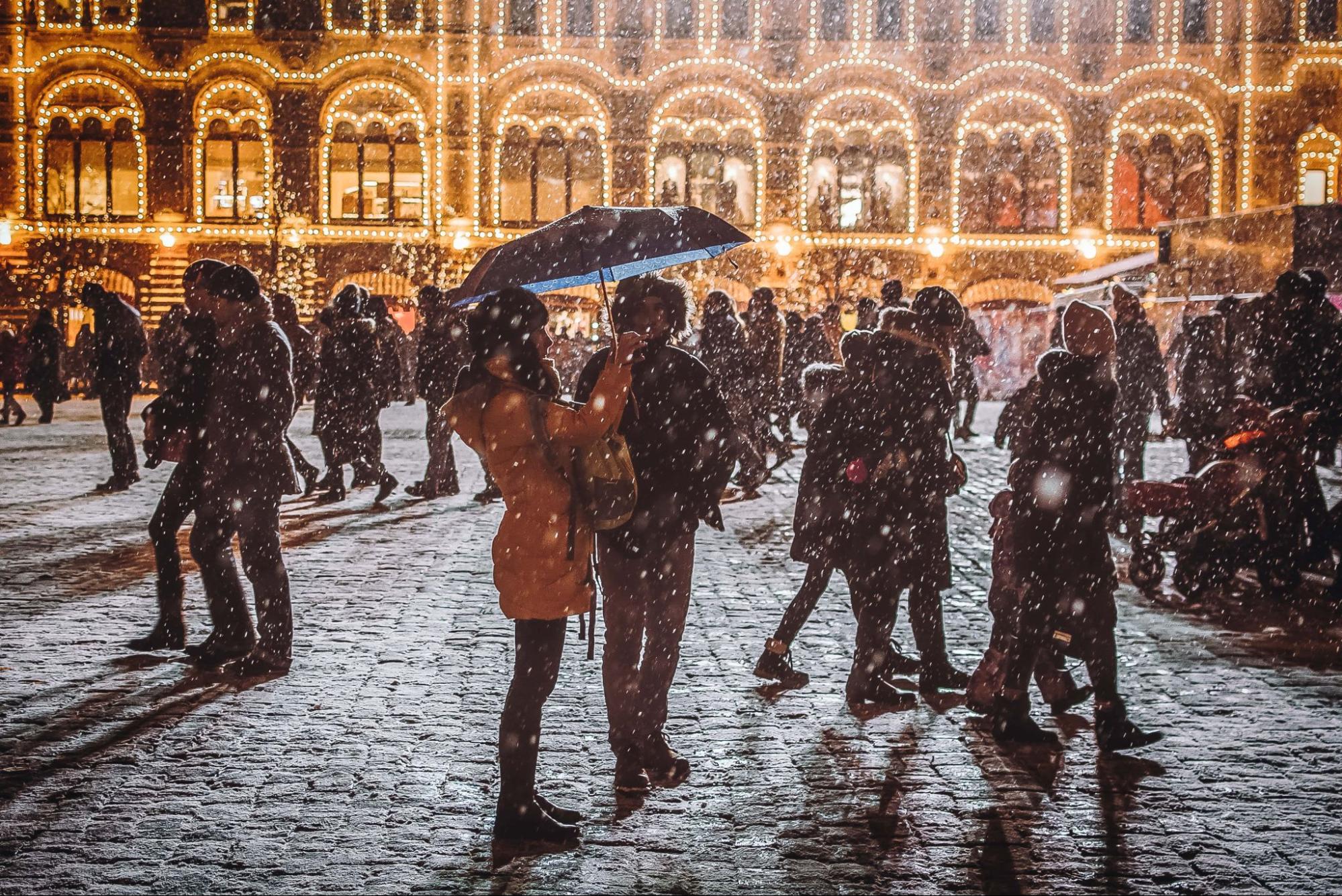 A young couple bundled up for the snowy weather in front of a beautifully lit building.