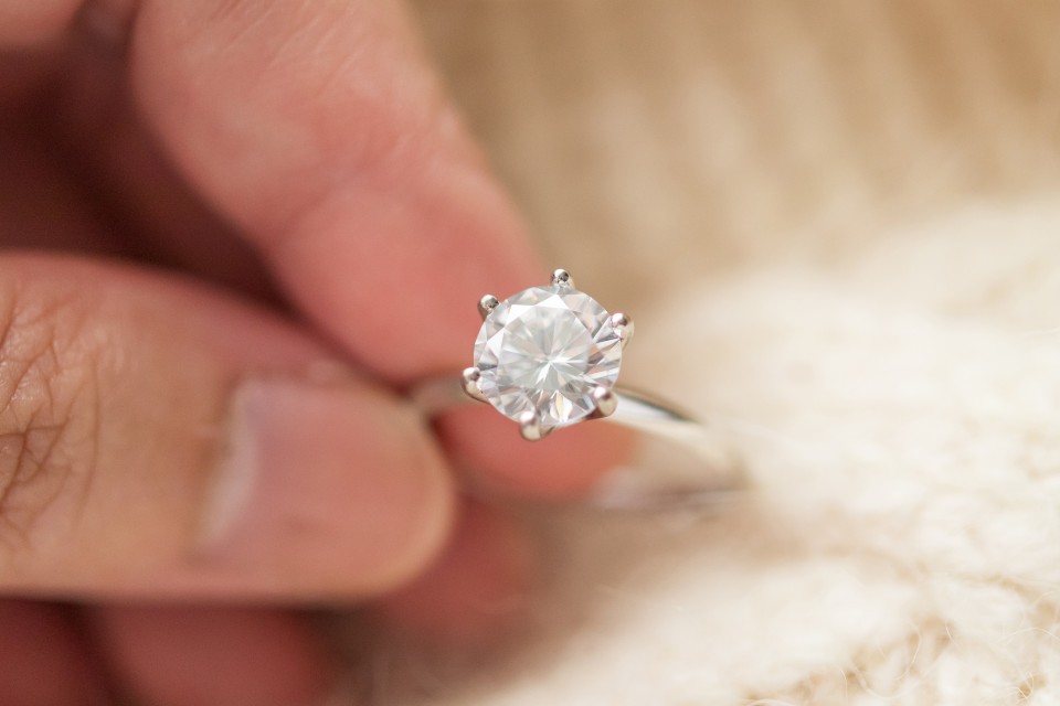 A close-up of a simple yet elegant solitaire engagement ring held between a person’s thumb and index finger over a marble backdrop.