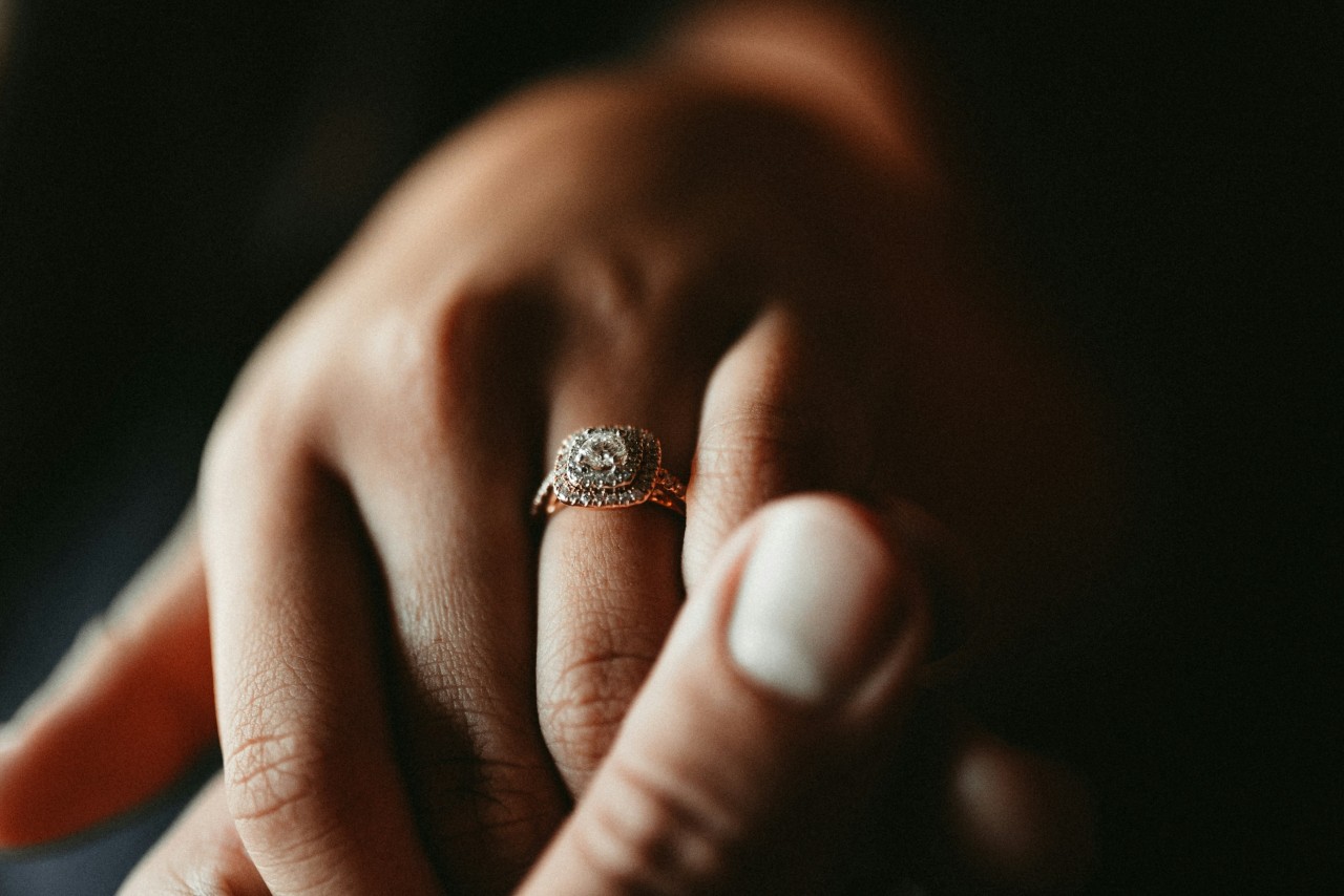 A close-up of a couple holding hands against a dark background, an elegant halo engagement ring visible on the bride-to-be’s finger.