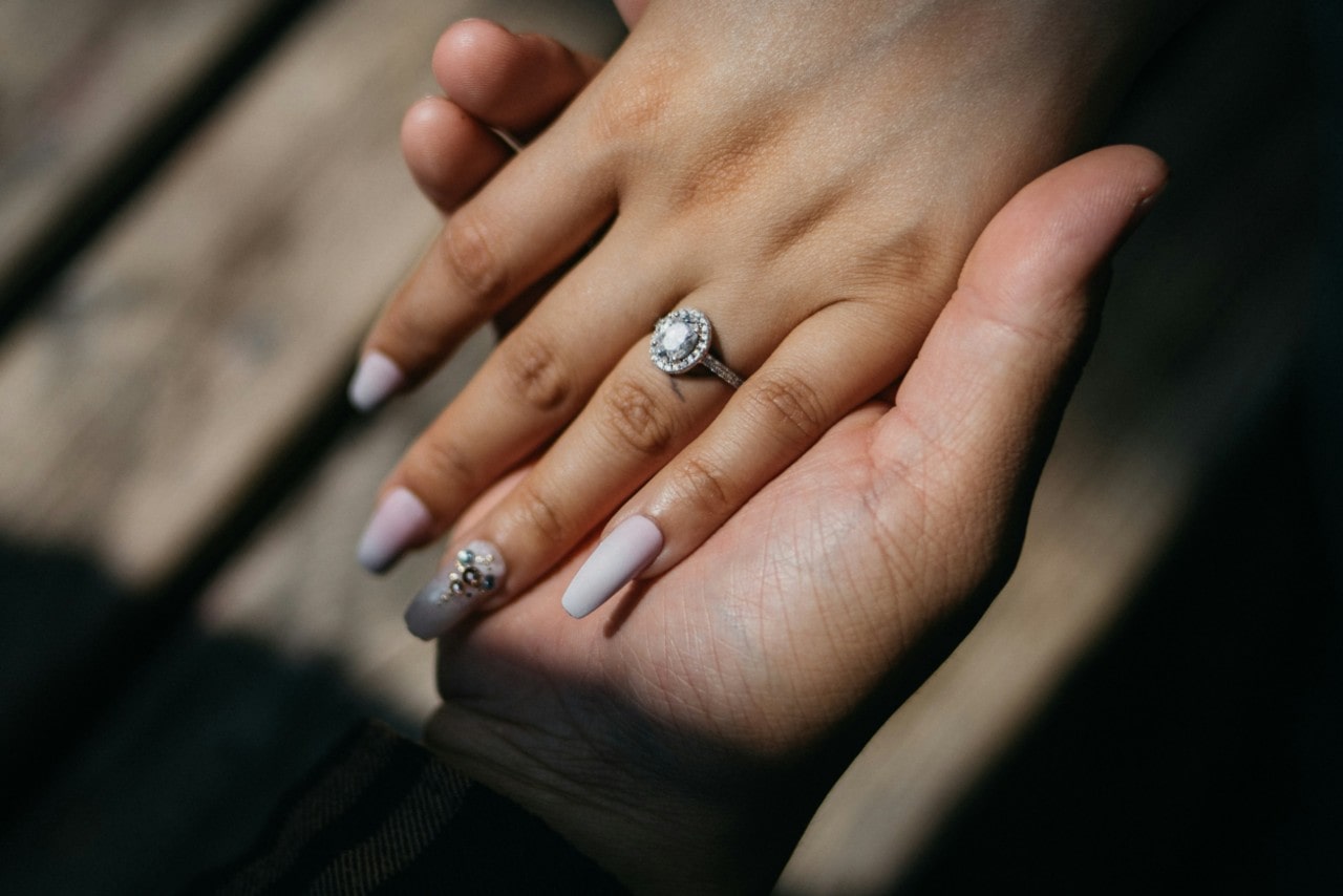 A close-up of a man’s hand holding his bride-to-be’s manicured hand, a dazzling engagement ring on her finger.