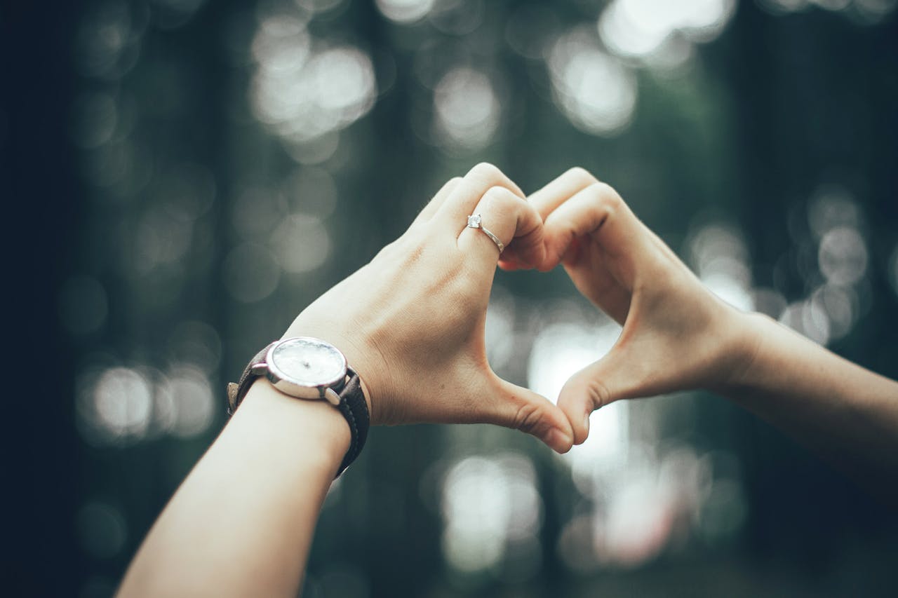 A close-up of a person’s hands, with an engagement ring on their finger, forming a heart shape with their fingers.