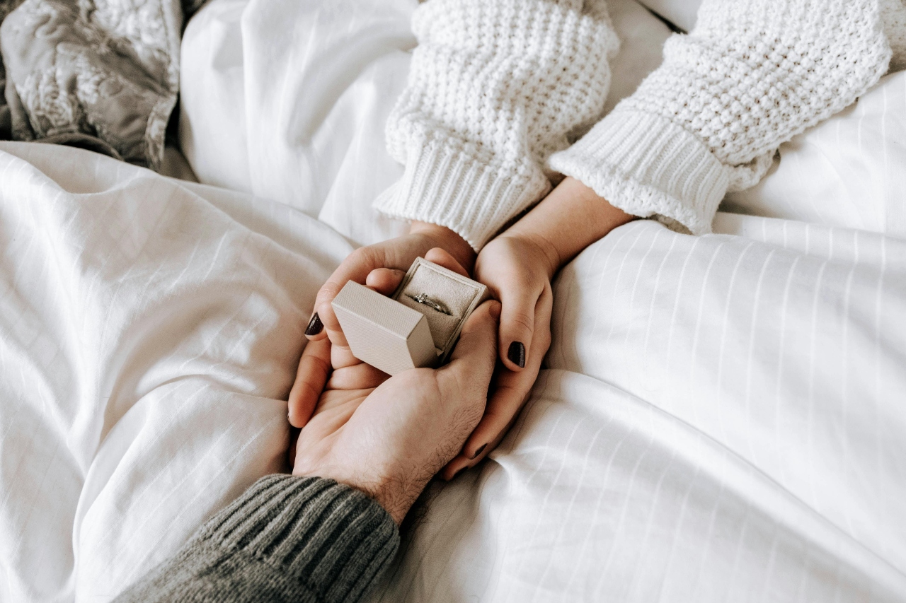 A comfortably dressed couple under a blanket, holding an engagement ring in their hands.