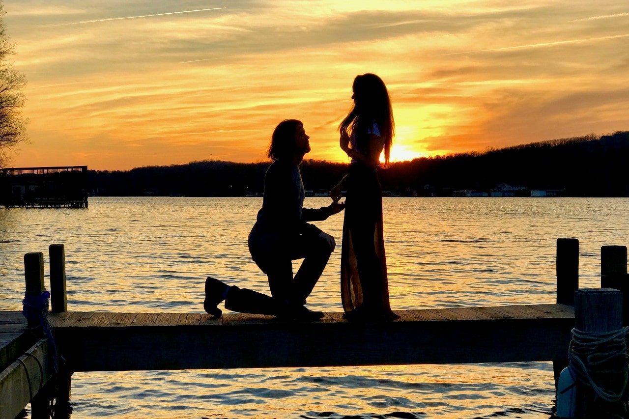 A man proposing to his bride-to-be on a picturesque waterfront at sunset.